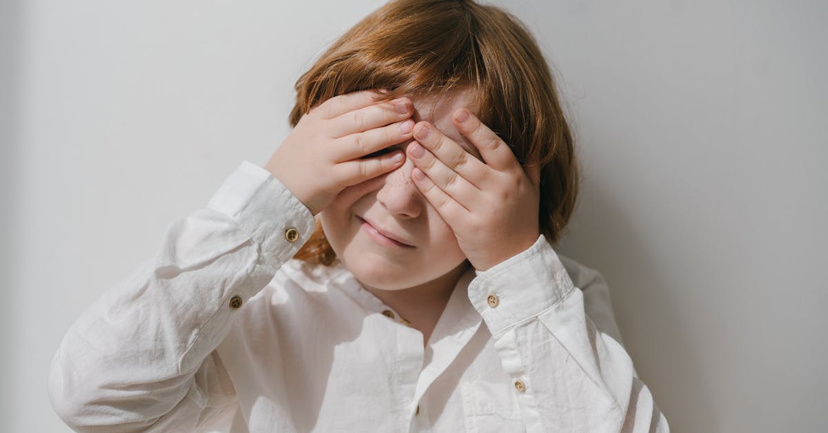 Caucasian child with red hair covers face with hands in bright indoor setting.